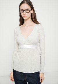 Ανοιχτό γκρι μπλούζα με μακριά μανίκια, λαιμόκοψη σε σχήμα V, σχεδιασμός τύπου ζακέτας και λευκή ζώνη από σατέν ως διακοσμητικό. Μαλακή υφή και άνετη εφαρμογή.