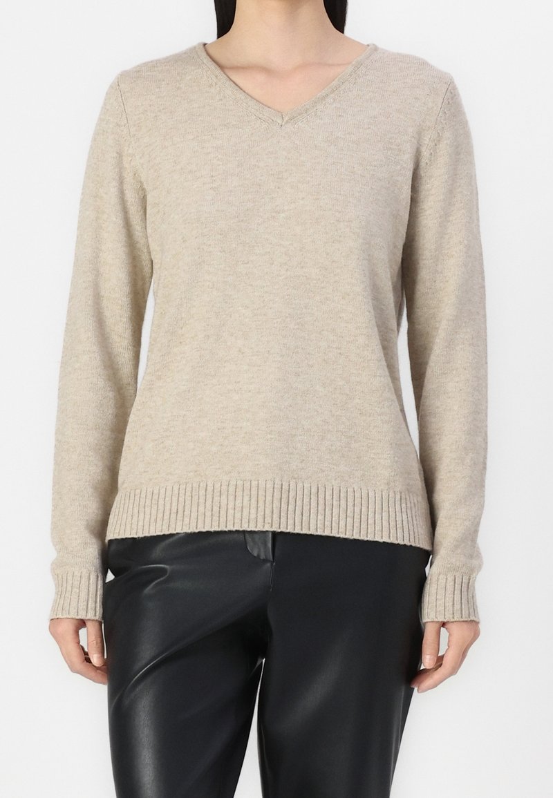 Pull beige à col en V et manches longues avec poignets et ourlet côtelés, porté avec un pantalon en cuir noir.