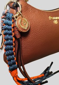 Borsa a mano in pelle marrone con trama, con emblema dorato a forma di "P" e portachiavi in paracord intrecciato blu e arancione attaccato a un anello dorato.