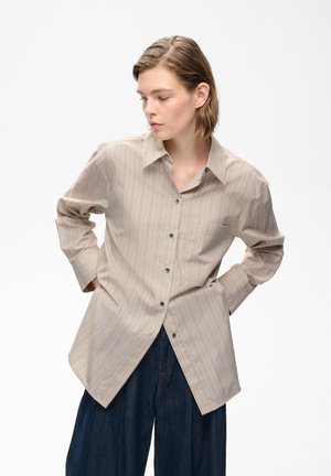 Junge Person trägt ein beige gestreiftes Langarmshirt und dunkle, weite Hose, steht mit den Händen in den Taschen und schaut nach links.