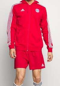 Un sweat à capuche rouge zippé avec des rayures blanches le long des manches, orné d'un logo brodé sur la poitrine, assorti à un short rouge.