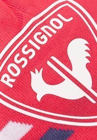Červená pletená tkanina s bílým logem stylizovaného kohouta, umístěného v oválném rámečku s textem "ROSSIGNOL," zdůrazňující texturu a design.
