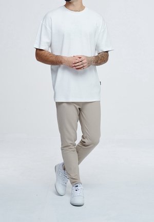 Mann mit Tätowierungen, der ein schlichtes weißes T-Shirt, beige Hose und weiße Sneaker trägt, steht mit überkreuzten Beinen und verschränkten Händen.