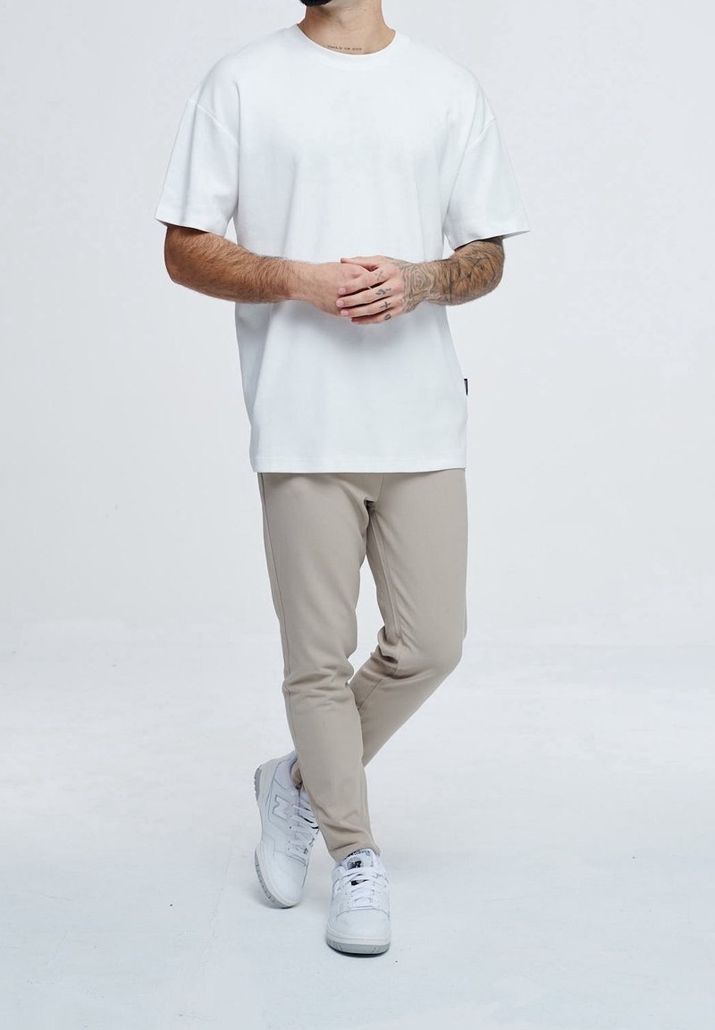 Homme avec des tatouages portant un t-shirt blanc uni, un pantalon beige et des baskets blanches, debout, jambes croisées et mains jointes.