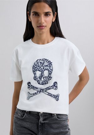 Mujer que lleva una camiseta blanca con un gráfico de una calavera y huevecillos cruzados, hecho de texto estilizado, combinado con unos vaqueros negros descoloridos.