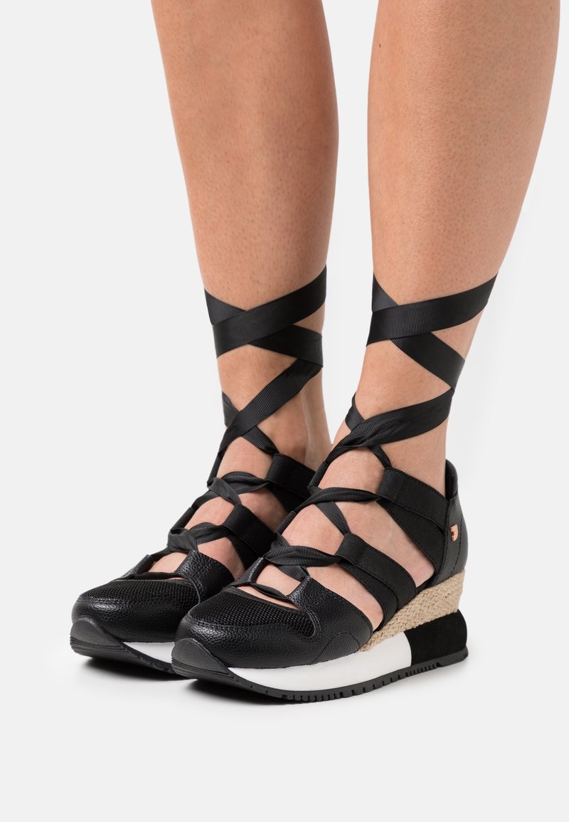 Pieds portant des sandales compensées noires avec des rubans croisés autour de la cheville et des semelles texturées sur fond blanc.