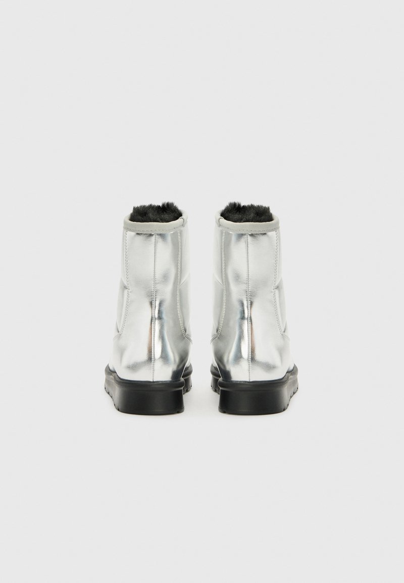 Une paire de bottes d'hiver métalliques argentées avec doublure en fourrure noire et semelles noires, vues de l'arrière sur un fond blanc uni.