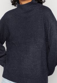 Mörkblå stickad tröja med hög ribbad krage, texturerat mönster och sänkt axel, med avslappnad passform och ribbade manschetter.