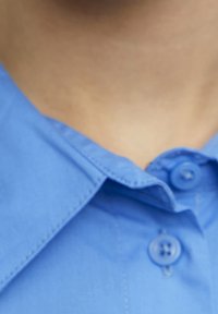 Niebieska koszula zapinana na guziki z wywiniętym kołnierzykiem, z dwoma guzikami w pobliżu dekoltu i gładką, teksturowaną tkaniną.