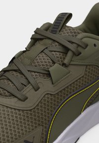 Κοντινό πλάνο αθλητικού παπουτσιού από ελαφρώς πράσινο δίχτυ, με κορδόνια, μαξιλαρωμένη γλώσσα και πλευρική ρίγα με κίτρινη λεπτομέρεια πάνω σε λευκή σόλα.