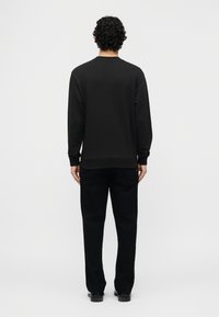 Άνδρας με κατσαρά μαύρα μαλλιά, φοράει μακρυμάνικο μαύρο πουλόβερ, μαύρο παντελόνι και μαύρα παπούτσια, στέκεται με την πλάτη γυρισμένη σε λευκό φόντο.