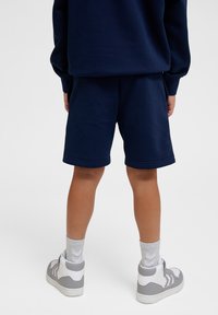 Navyblauwe katoenen shorts met een elastische tailleband, gecombineerd met grijze high-top sneakers en lichtgrijze sokken, bekeken vanuit de achterkant.