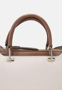 Sac à main avec corps blanc texturé et anses en cuir marron attachées par une quincaillerie métallique argentée, avec une fermeture éclair sur le dessus.