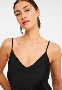 Schwarzes Trägershirt mit dünnen Trägern, das über einen tiefen V-Ausschnitt und eine glatte, matte Textur verfügt. Kombiniert mit fallenden, mit Perlen besetzten Ohrringen.