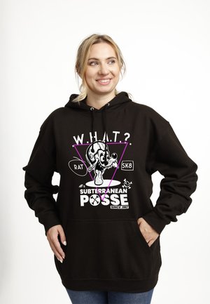 Kobieta uśmiechająca się w czarnej bluzie z kapturem z kreskówkową szczurą w hełmie, fioletowym trójkątem i napisem "W.H.A.T.? Subterranean Posse Since 1967."