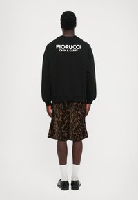 Muž stojaci chrbtom, oblečený v čiernej mikine s nápisom "FIORUCCI CASH & CARRY" a vzorovaných hnedých šortkách, bielych ponožkách a čiernych topánkach.