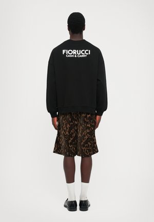 Mann som står med ryggen til, iført svart sweatshirt med teksten "FIORUCCI CASH & CARRY" og mønstrede brune shorts, hvite sokker og svarte sko.