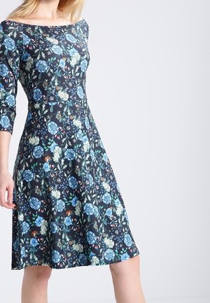 Frau trägt ein knielanges schwarzes Kleid mit blauen und weißen Blumenmustern und Dreiviertelärmeln vor einem schlichten hellen Hintergrund.
