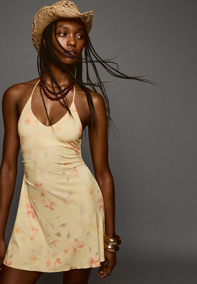 Robe florale jaune clair avec un décolleté en V et des bretelles halter. Accentuée de bracelets en or et d'un chapeau de cowboy en paille. Texture lisse et design fluide.