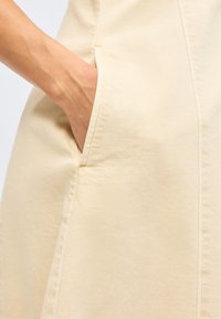 Χέρι που έχει τοποθετηθεί στην πλαϊνή τσέπη ενός ρούχου από κρεμ υφασμάτινο ύφασμα με υφή, με ορατές ραφές και λεπτομέρειες ραφών.