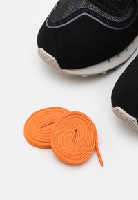 Schwarzer Sneaker mit einer strukturierten Oberseite und grauen Akzenten, kombiniert mit zwei aufgerollten orangefarbenen Schnürsenkeln auf einem weißen Hintergrund.
