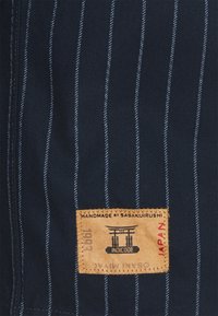 Navy fabric with thin light blue vertical stripes, featuring a sewn tan label with black text and a graphic, indicating handmade origin from Japan.