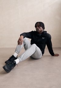 Svart zip-up hettegenser med EA7-logo, lysegrå avsmalnede bukser og svarte sneakers. Avslappet posering på et lyse farget gulv.