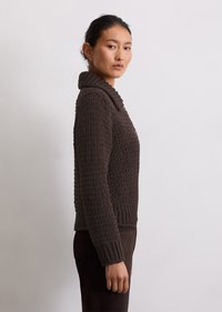Maglione marrone lavorato a maglia con colletto alto, motivo texturizzato, polsini a coste e vita aderente. Il modello lo indossa con pantaloni marrone scuro.
