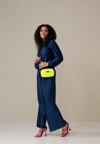 Navyblaues, plissiertes Jumpsuit, pinkfarbene Riemchenheels und eine kleine strukturierte, neongelbe Umhängetasche. Glatte Textur und lockere Passform.