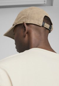 Șapcă de corduroy de culoare bej cu vizieră arcuită și curea ajustabilă la spate. Are o etichetă mică cu logo pe curea. Purtați-o cu o cămașă bej.