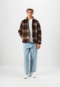 Young man wearing a brown plaid jacket, white t-shirt, light blue polka dot jeans, white sneakers, and a beige cap standing against a white wall.