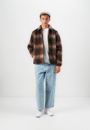 Jonge man in een bruine geruite jas, wit T-shirt, lichtblauwe spijkerbroek met stippen, witte sneakers en een beige pet, staand tegen een witte muur.