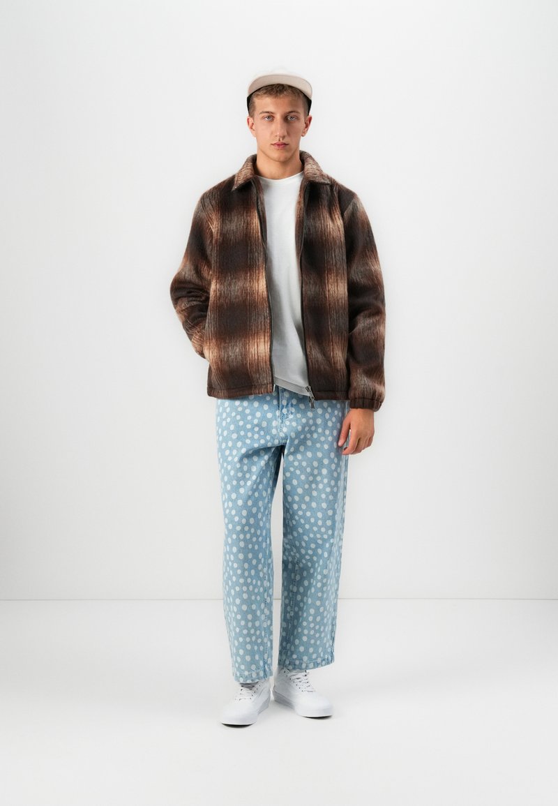 Young man wearing a brown plaid jacket, white t-shirt, light blue polka dot jeans, white sneakers, and a beige cap standing against a white wall.