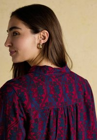 Czerwona i niebieska bluzka w kwiatowy wzór z marszczonym tyłem. Tkanina ma subtelnie wyczuwalną fakturę. Złote kolczyki w kształcie kół uzupełniają całość.