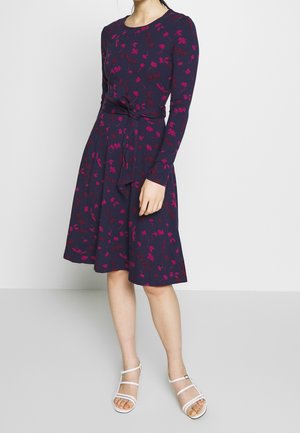 Femme portant une robe bleu marine mi-longue à manches longues avec des imprimés floraux roses, associée à des sandales blanches à lanières.