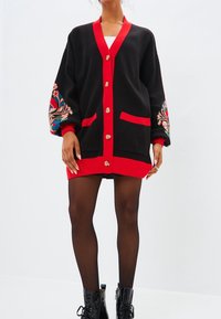 Black oversized cardigan with red accents, featuring large buttons, two front pockets, and colorful floral embroidery on sleeves.