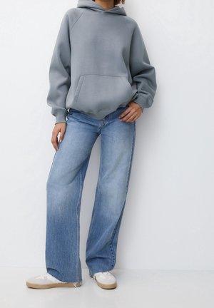 Person iført en grå oversized hættetrøje, lyseblå vidde jeans og hvide sneakers, der står mod en ensfarvet hvid baggrund.