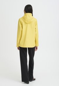 Veste imperméable jaune avec capuche, manches longues et coupe décontractée, associée à un pantalon large noir, vue de dos.