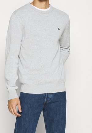 Mand iført en lysegrå crewneck-trøje med lille grønt krokodillelogo på brystet og blå jeans, står med venstre hånd afslappet.