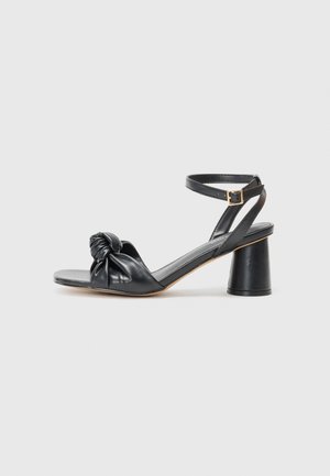 Sandales à talons en cuir noir avec un design de devant noué, bout ouvert et bride à la cheville ; elles présentent un talon chunky et cylindrique.