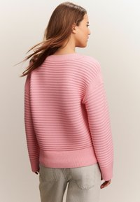 Kvinna med långt brunt hår som bär en ribbad rosa tröja och ljusgrå jeans, vänd bort mot en enkel ljus bakgrund.