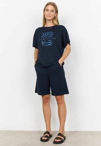 Námořnická modrá bavlněná tričko s oversized střihem a krátkými rukávy, s výrazným světle modrým grafickým textem a abstraktním designem listu. V kombinaci s odpovídajícími šortkami.