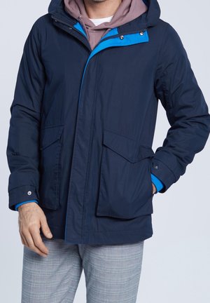 Hombre con chaqueta con capucha azul marino sobre sudadera con capucha malva y pantalones de cuadros grises, con una mano en el bolsillo de la chaqueta.