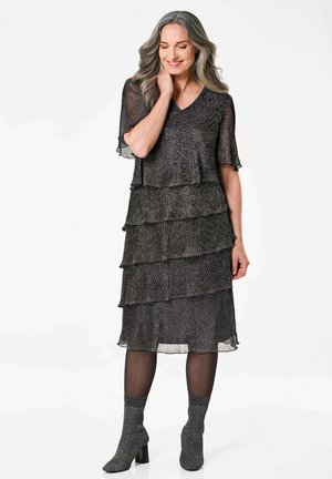 Schwarzes, schichtartiges Kleid aus strukturiertem Stoff mit V-Ausschnitt und kurzen Ärmeln. Kombiniert mit grauen Knöchelstiefeln und schwarzen Strumpfhosen.