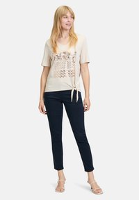 Beige kortærmet top med en sløjfe i siden, prydet med et metallisk blomsterprint. Parret med mørkeblå slim-fit bukser og åbne sandaler.