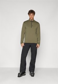 Camiseta de manga larga verde oliva con cremallera y cuello alto, combinada con pantalones negros y botas de esquí. Tejido suave con un diseño ajustado.