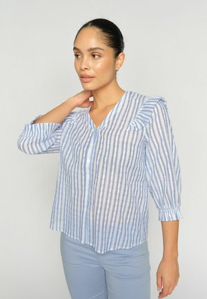 Woman with dark hair in a bun wears a light blue and white vertical striped blouse with ruffle details and light blue pants, against a plain background.
