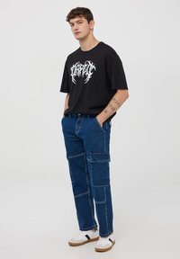 Czarna bawełniana koszulka z białym nadrukiem graficznym, połączona z niebieskimi spodniami cargo z denimu z wieloma kieszeniami i luźnym krojem. Białe sneakersy.