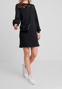 Černé šaty s dlouhými rukávy a krajkovým akcentem na výstřihu, doplněné malou černou crossbody taškou a bílými botami. Hladká textura.
