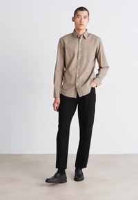 Camisa de botones marrón claro con cuello, combinada con pantalones negros y zapatos negros, de pie sobre un fondo neutro.